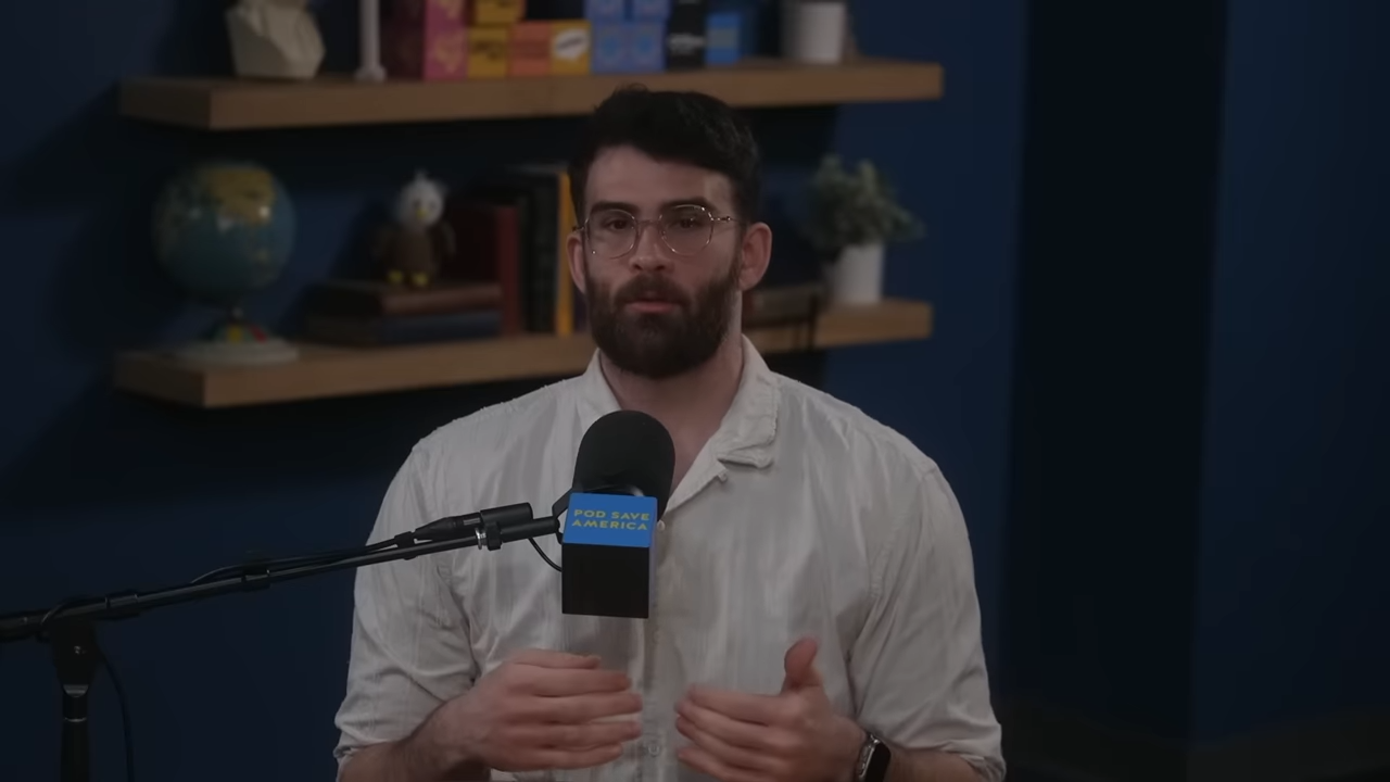 A man sits behind a microphone, hands raised to make a point