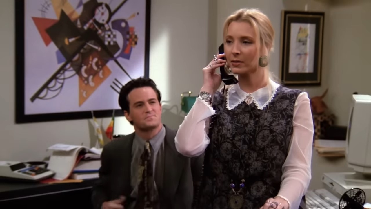 A woman sits on a desk talking on the phone while a man watches admirably 