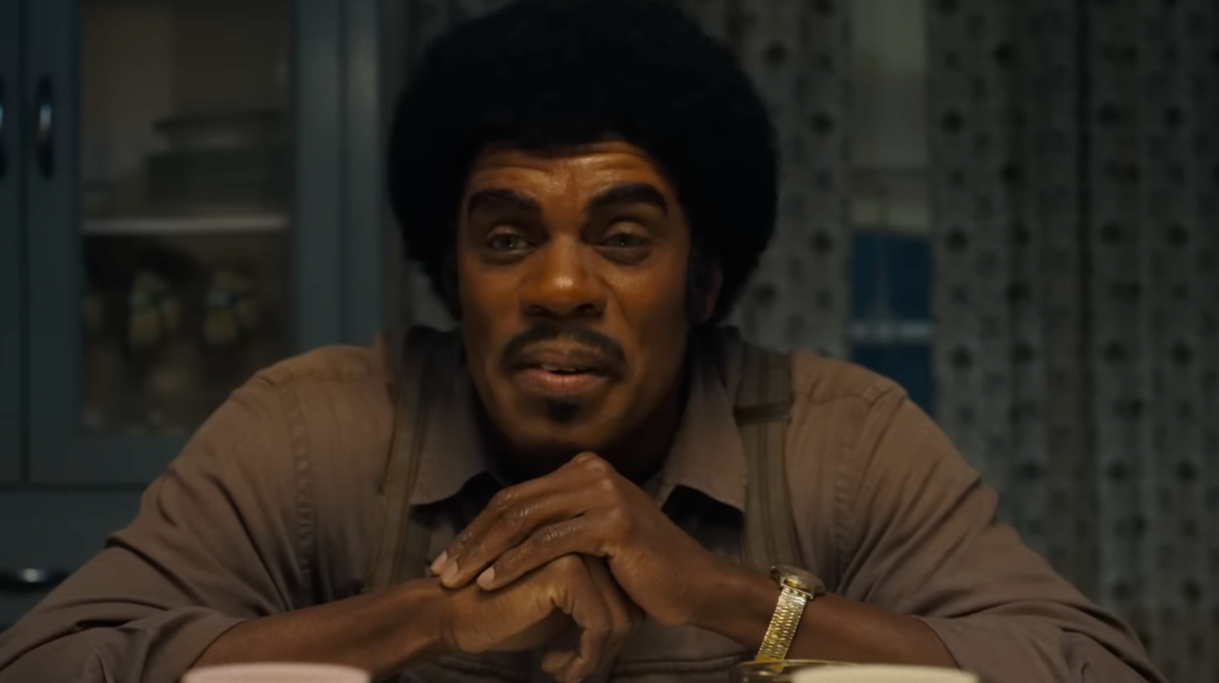 A man with an afro and a mustache rests his chin on his hands. The sensation is ominous.