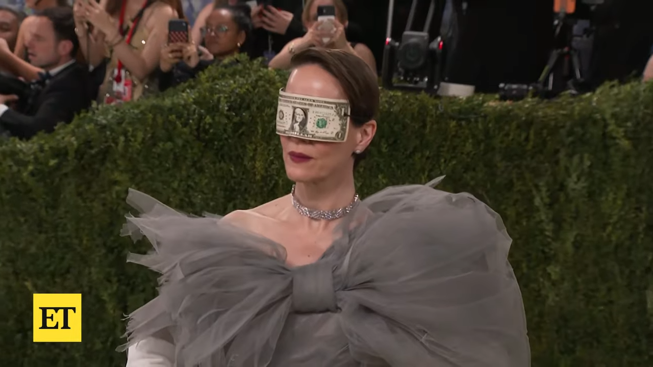An elegant woman stands at a formal event, blindfolded with dollar bill