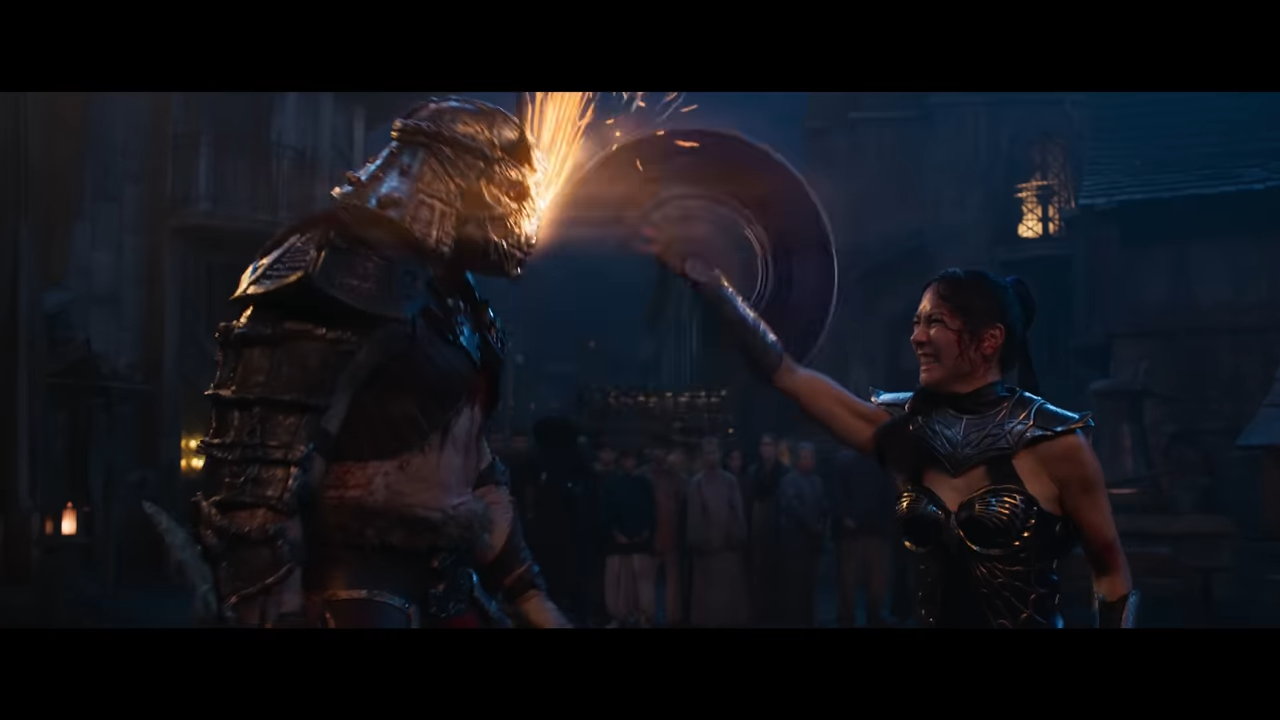 A woman holds a spinning metal blade to a tall, helmeted man's face, gritting her teeth as sparks fly