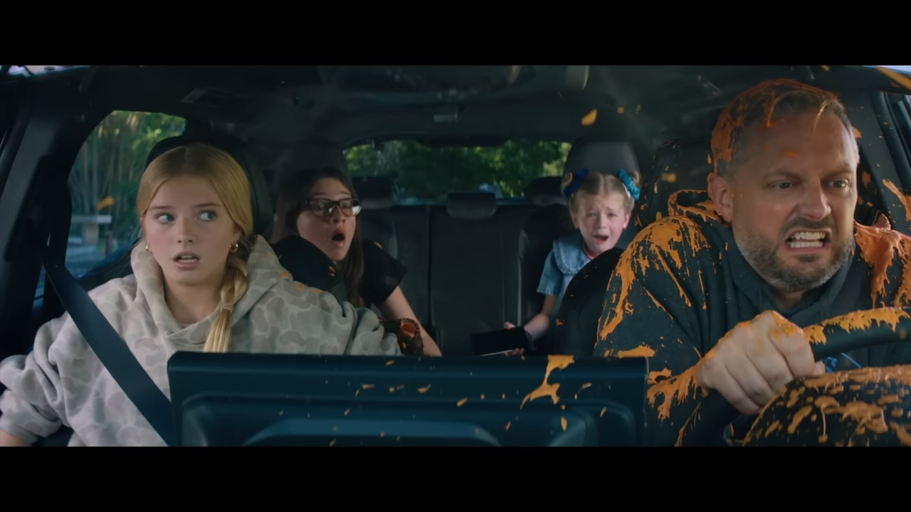 We see through the windshield a shocked family, the dad driving is covered in an orange smoothie
