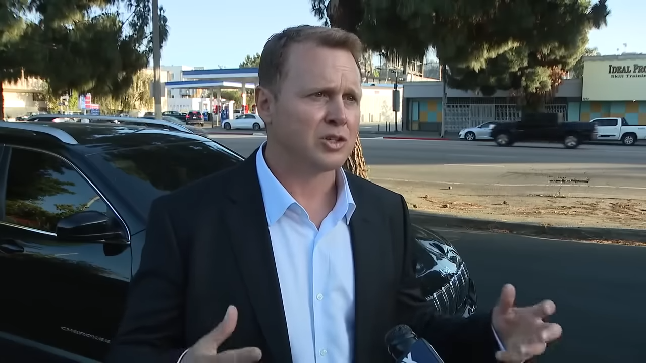 A man in an open collar shirt and sport jacket stands outside for a street interview
