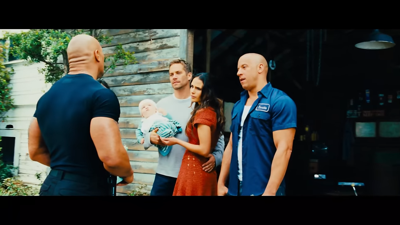 A couple with a baby stands beside a man in work shirt outside a garage
