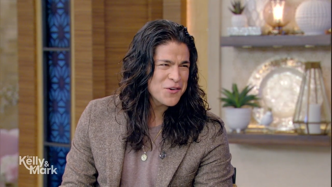 A man with long hair wearing a sports jacket sits for a warm TV interview