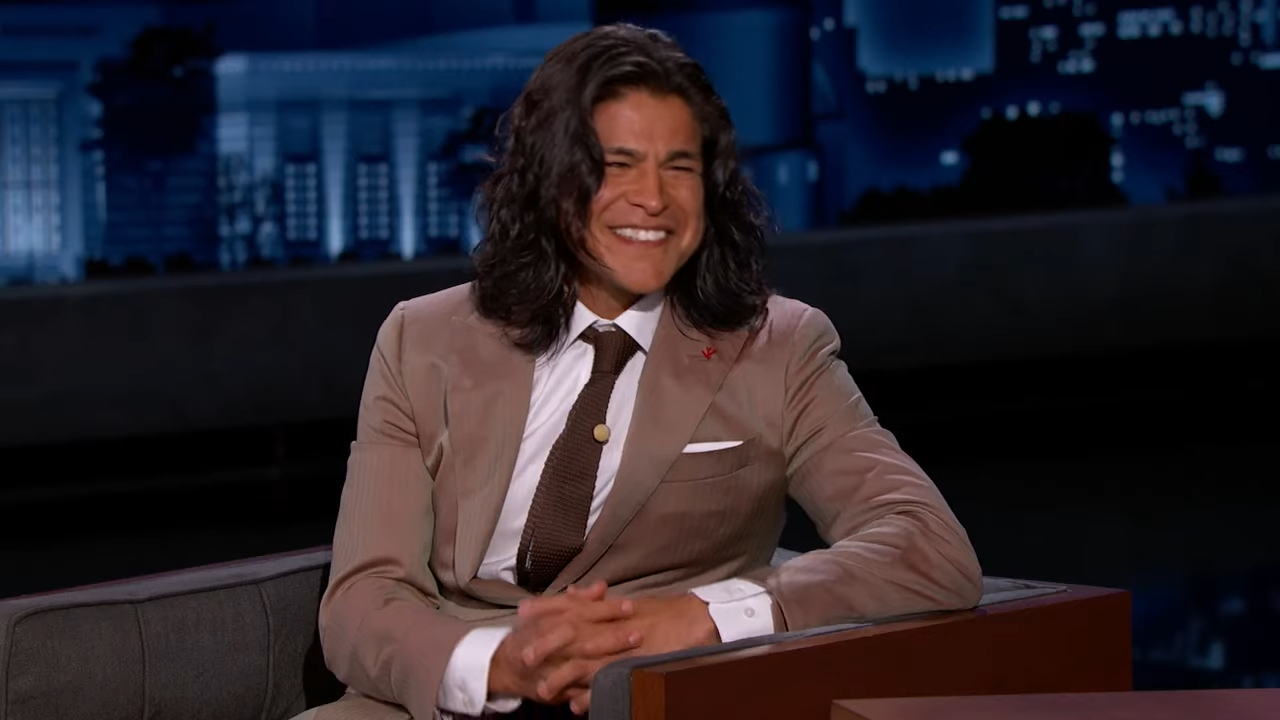 A man with long hair wearing a suit and tie leans on one arm and smiles during a late-night TV interview
