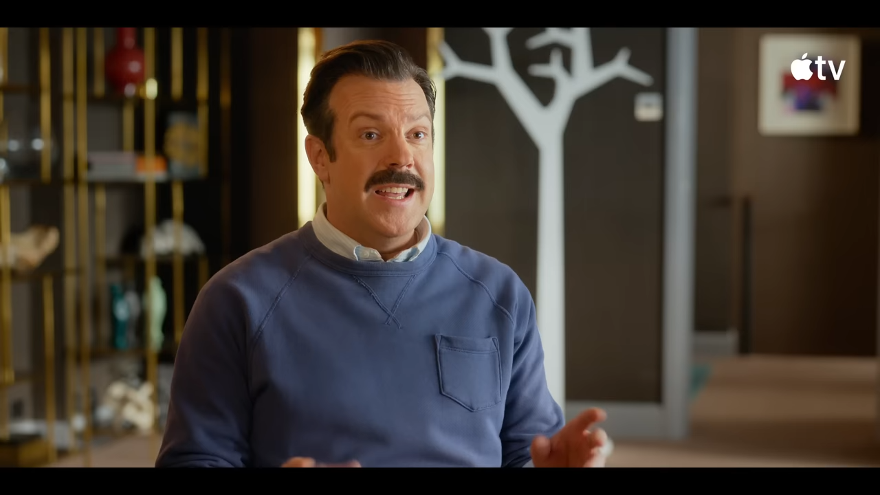 A mustached man in a sweater enthusiastically explains