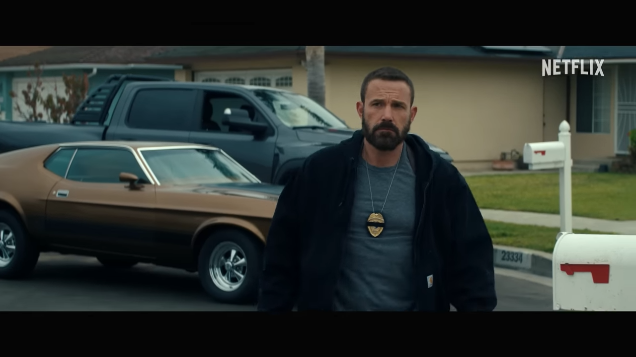 A man with a police badge around his neck walks through a suburban neighborhood. A vintage car is parked behind him.