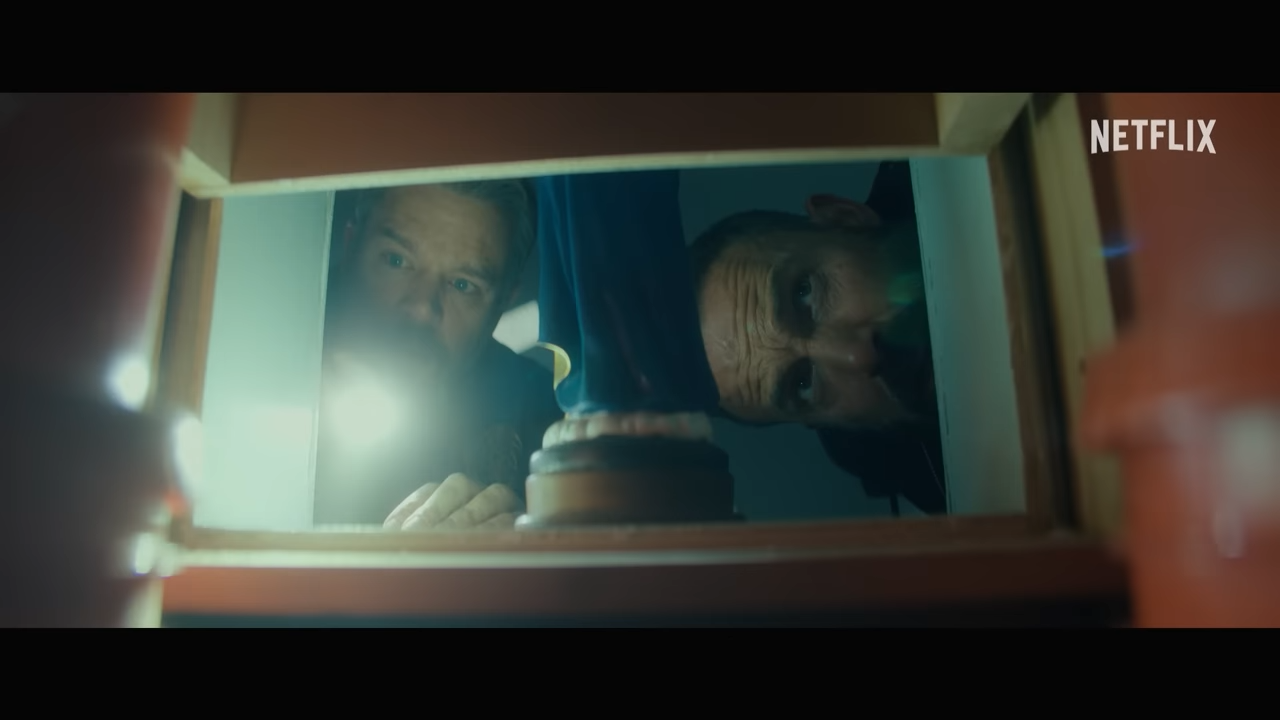 Two men use a flashlight to peer through a square cut hole in a wall