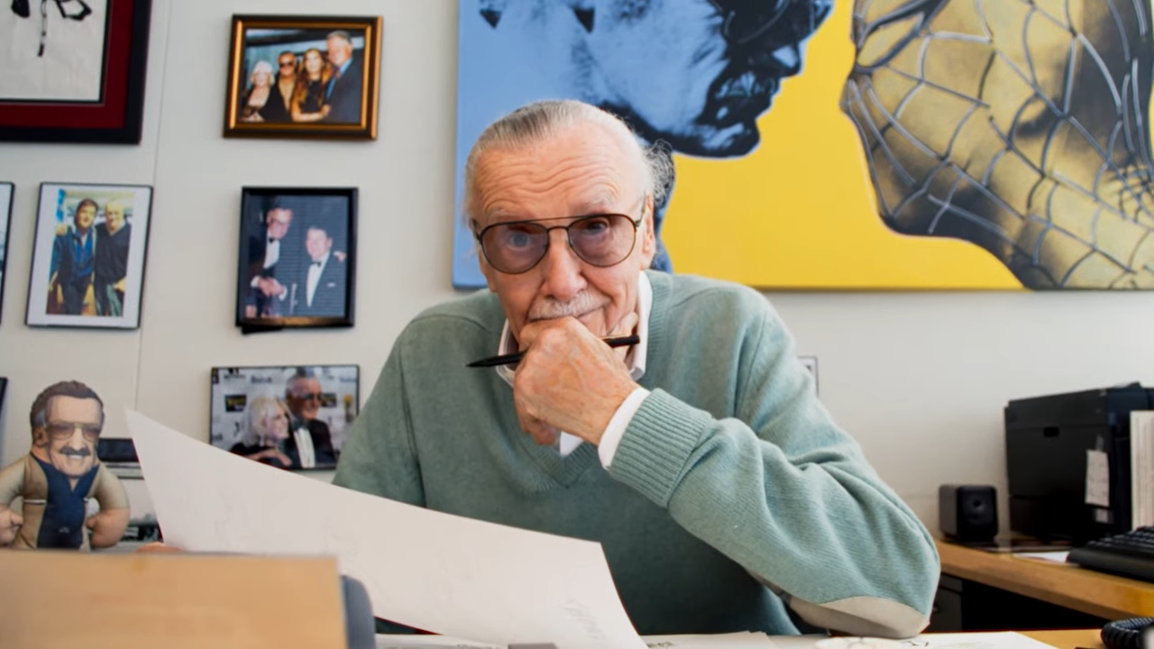 (50) Introducing Stan Lee on ElevenLabs 00-01-13 An AI generated image of Stan Lee seated at a desk. His hand is lifted to his chin as he smiles at the viewer