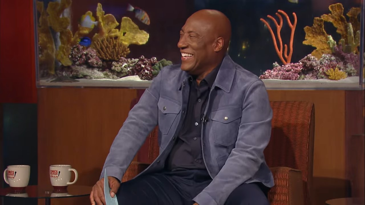 A man sits on a TV comedy talk show in front of a fish tank, laughing