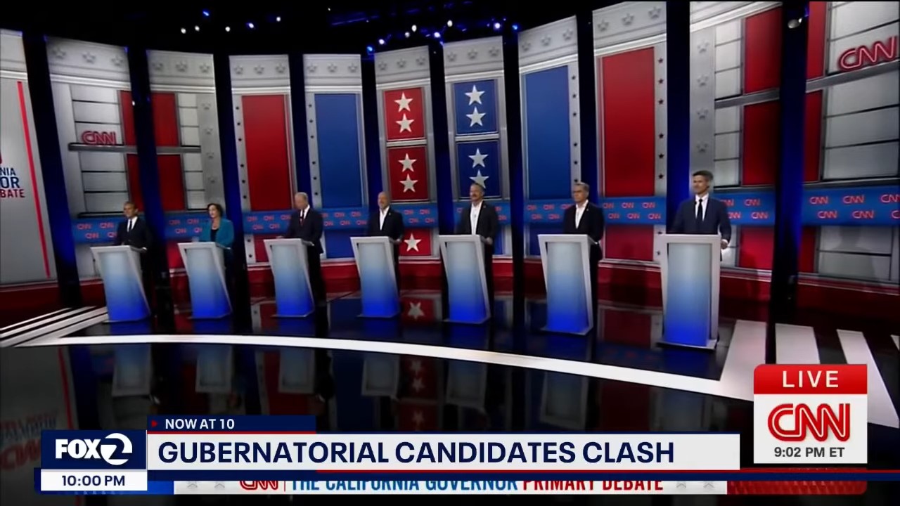 A televised debate stage with candidates at seven podiums