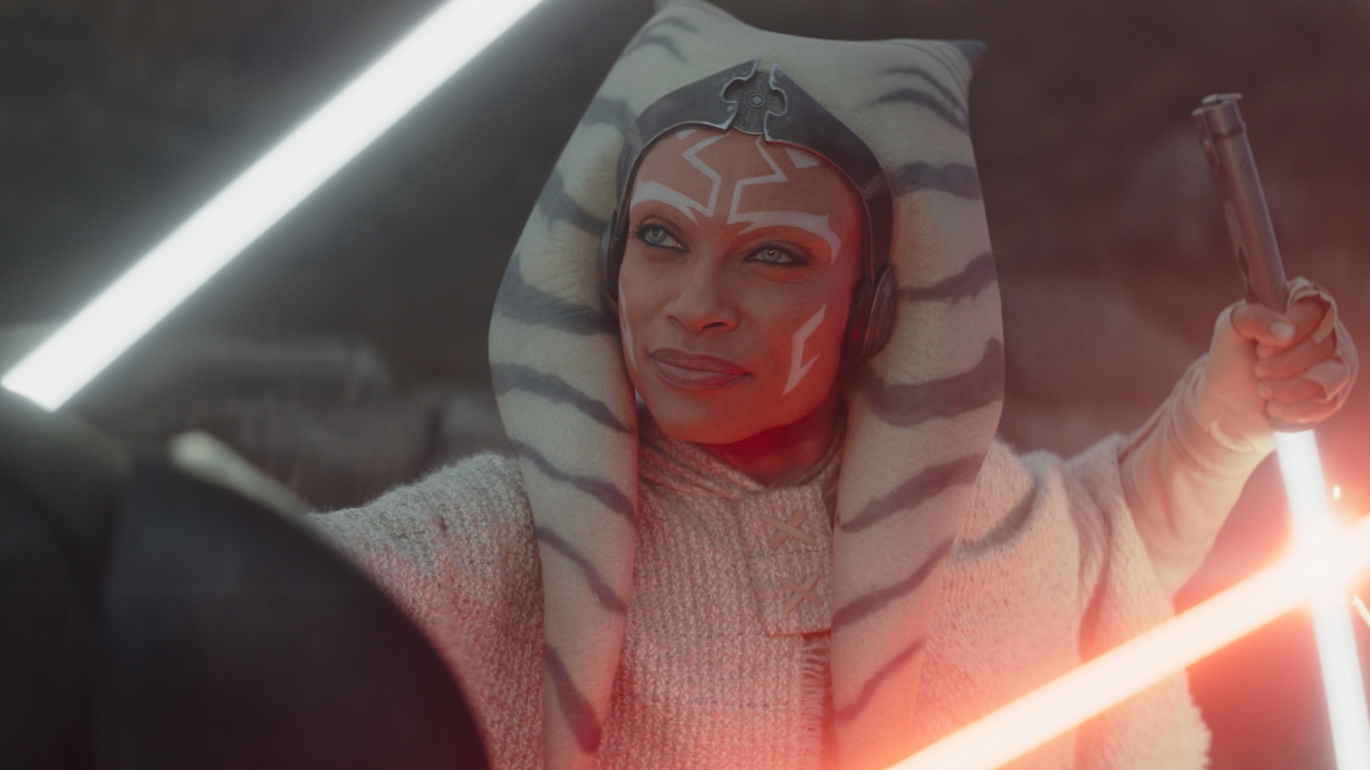 A woman with a head covering and face tattoos holds a light sabre