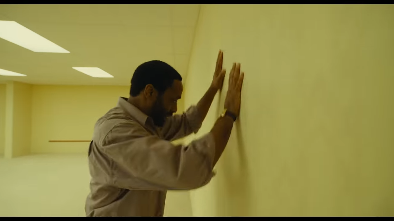 A man desperately presses his palms against a wall in an empty office space with sickly light