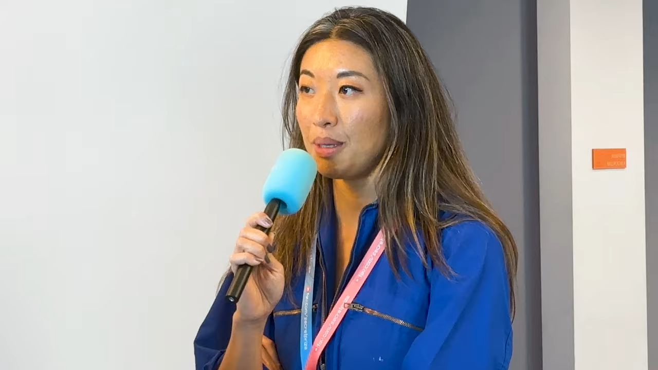 A woman holds a microphone, speaking against a blank wall