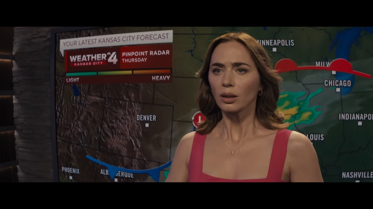 A woman stands in front of a TV weather map with a strange, unsettling look on her face