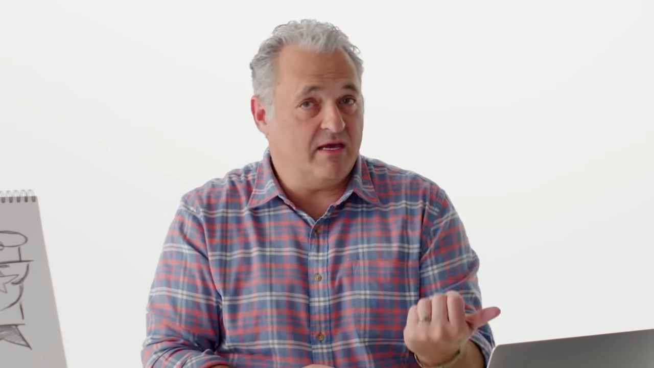 An older man in a button down shirt speaks directly into the camera, friendly and personable