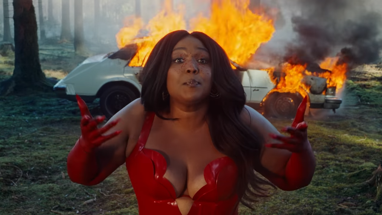A woman stands, unconcerned, in front of a burning car.