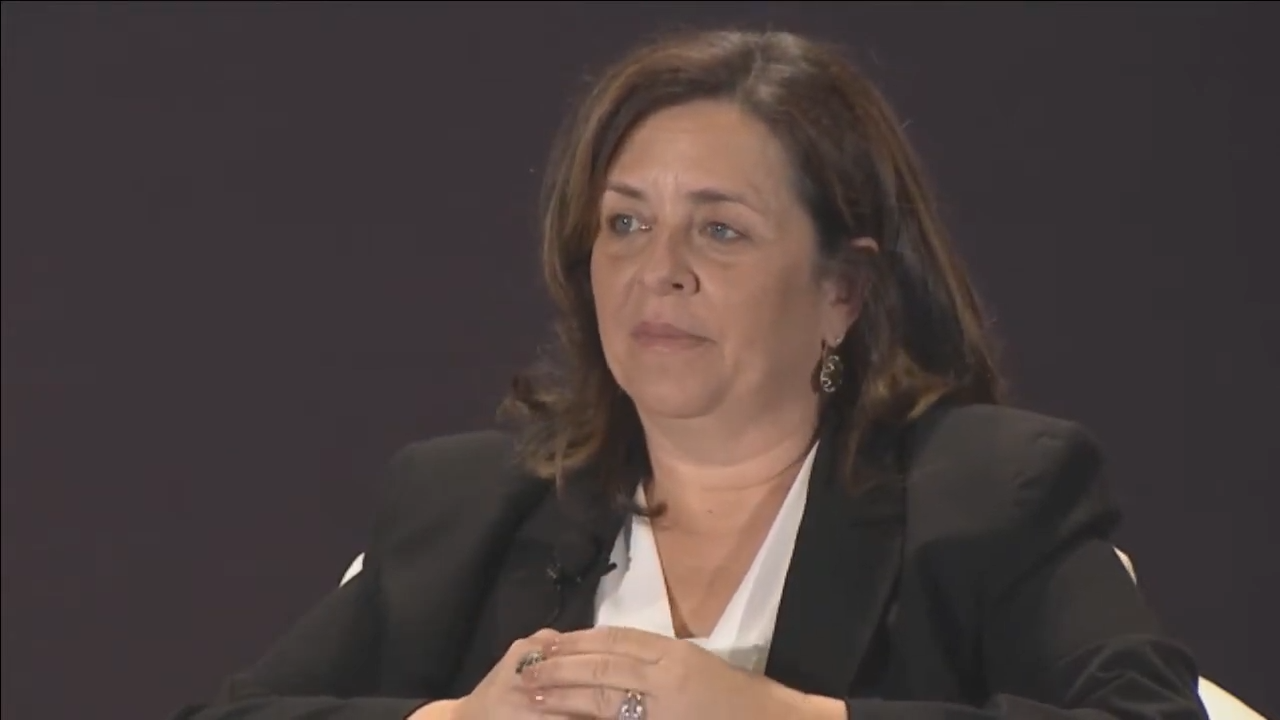 A woman in a business suit sits, hands folded, serious
