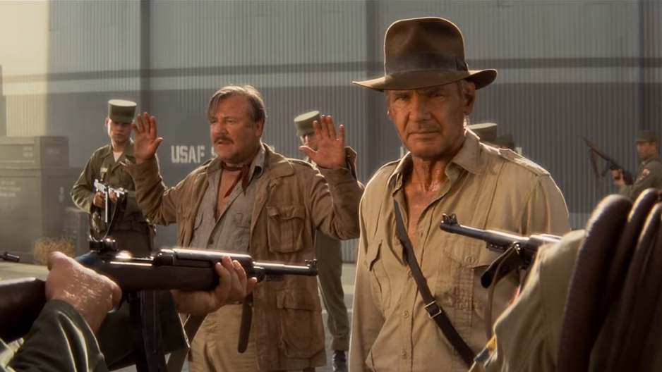 Indiana Jones and frightened man are surrounded by men pointing guns
