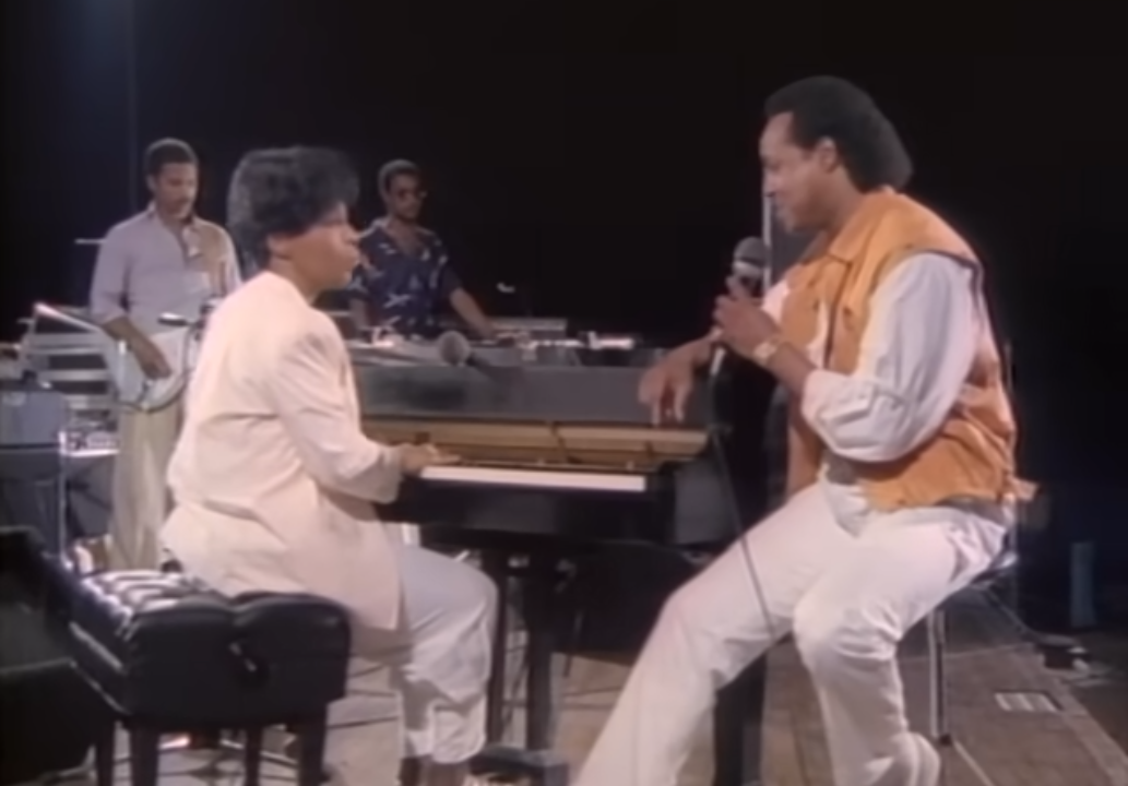 Woman dressed in white playing piano and singing with a man in orange and white