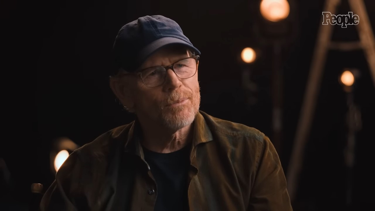 Ron Howard in glasses and a ballcap sits in a dark studio for an interview