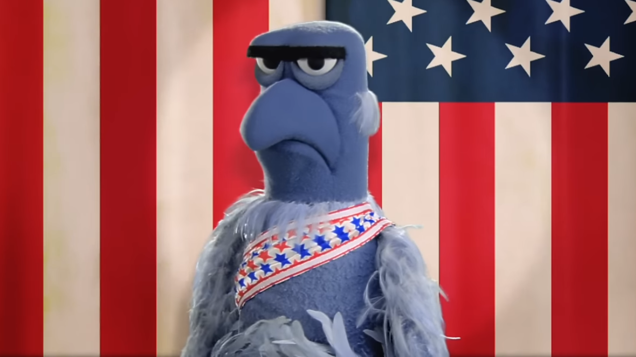 Sam Eagle looking sternly at the camera with a patriotic sash, in front of the American flag
