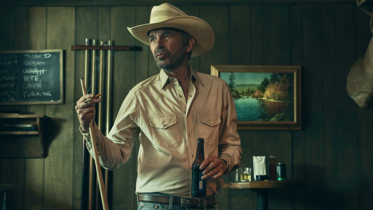 A grizzled cowboy stands in a bar, pool cue in one hand and a beer in the other