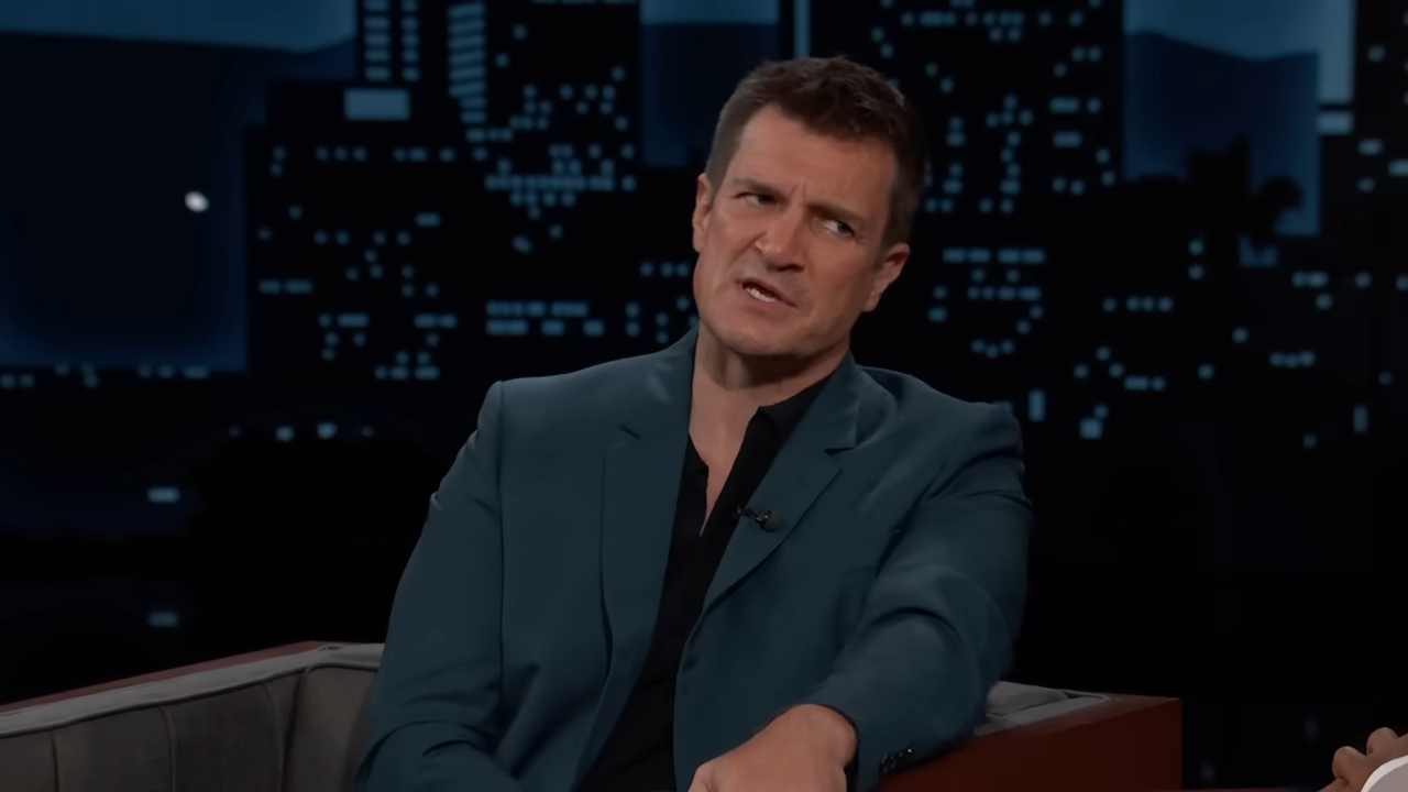 Nathan Fillion leans toward the host on a late-night show, making a semi-serious face