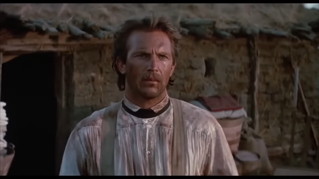 A stern-faced frontiersman stands, dirty and tired, outside his cabin