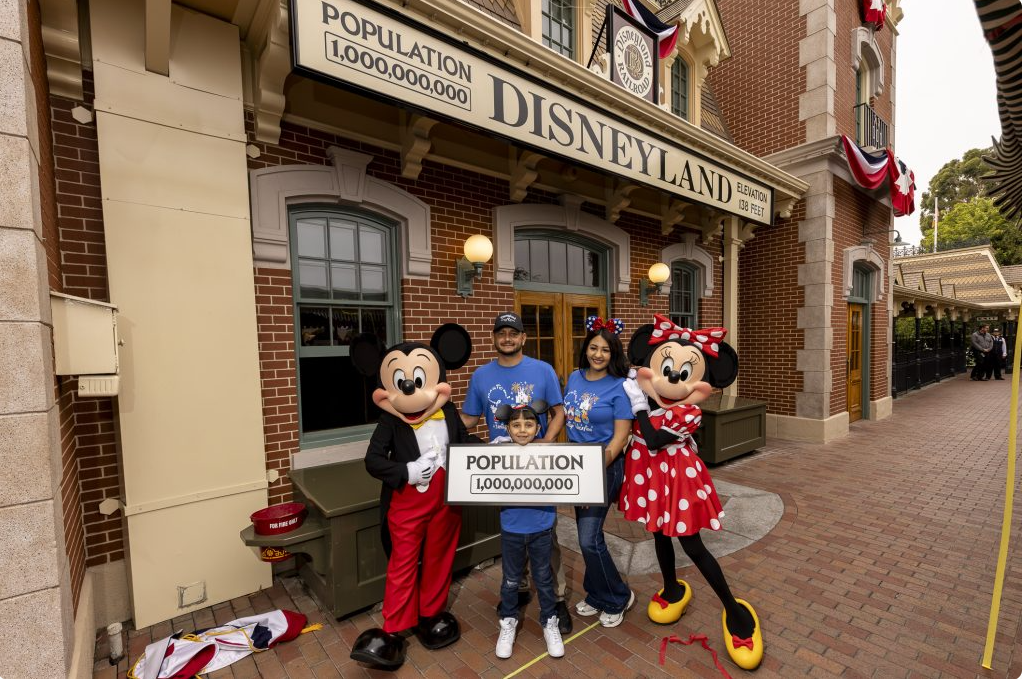Image of man, woman, and child holding a sign that says "Population 1,000,000,000" between Mickey and Minnie Mouse