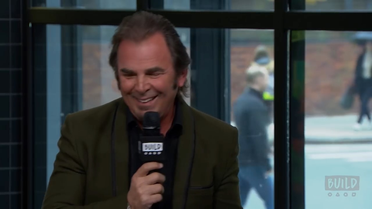 Jonathan Cain holds a microphone and smiles during a promotional interview