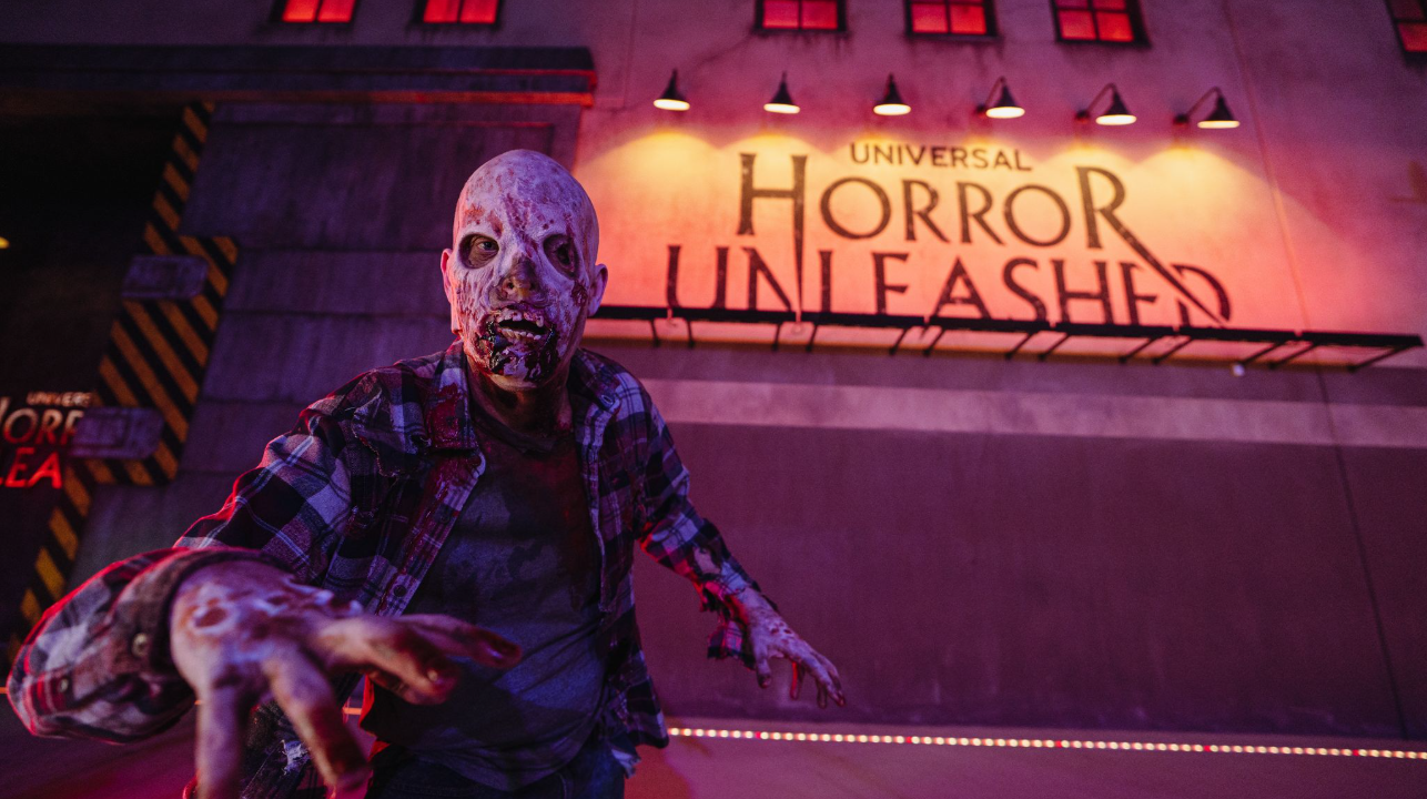 Universal Horror Unleashed Bloodied, disfigured man reaches for camera in dark alley with "Universal Horror Unleased" on a lit sign behind him