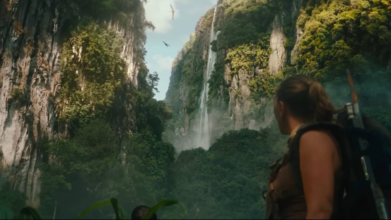 A woman looks up at a waterfall as flying dinosaurs circle