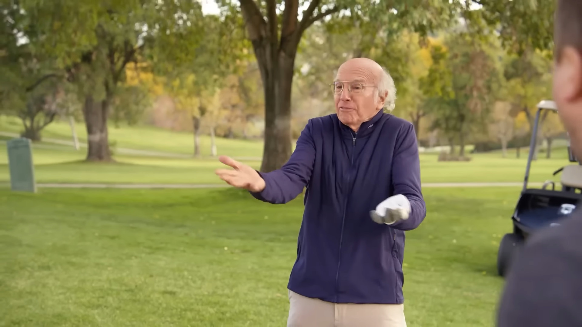 Larry David on a golf course, hands raised as if making an excuse