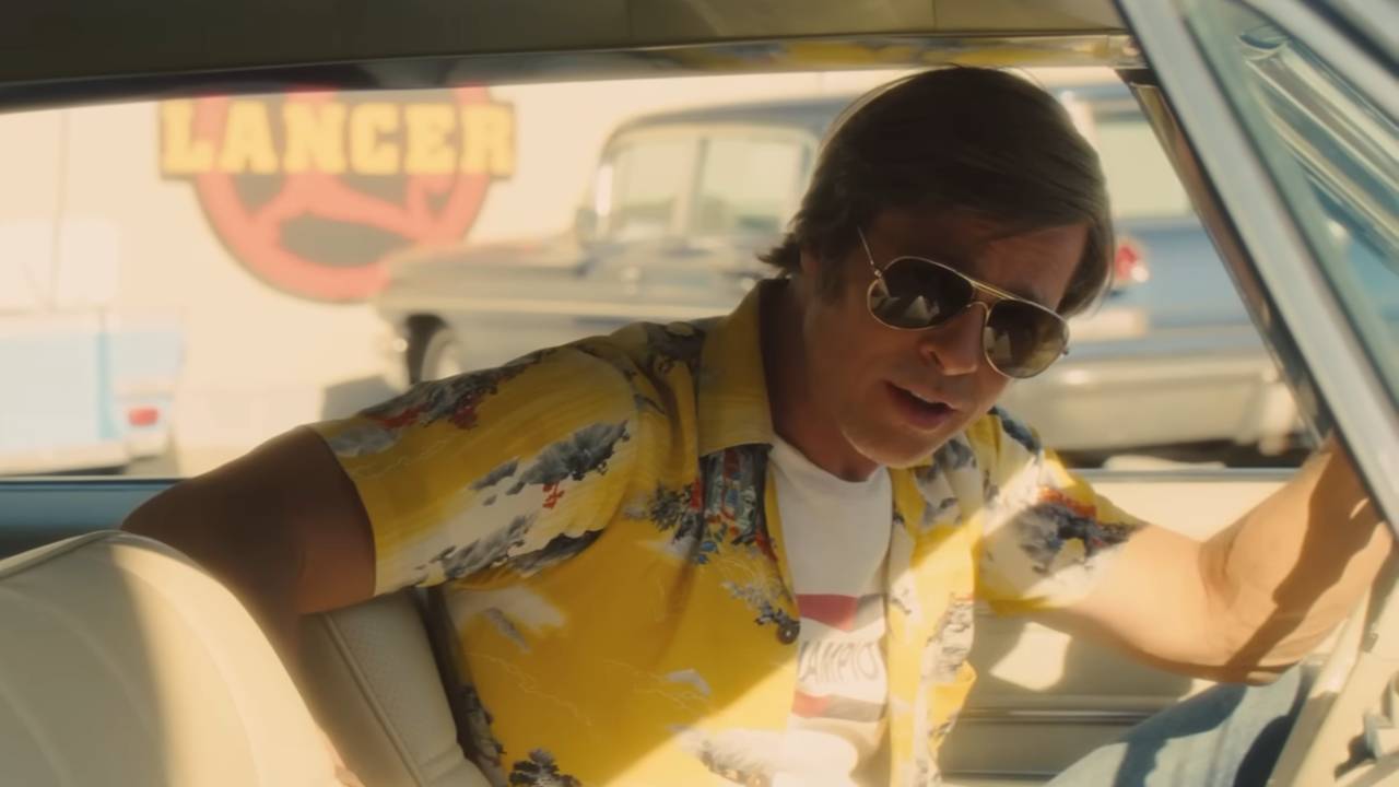 Brad Pitt as Cliff Booth A man in sunglasses sits behind the wheel of a car, looking through the passenger window