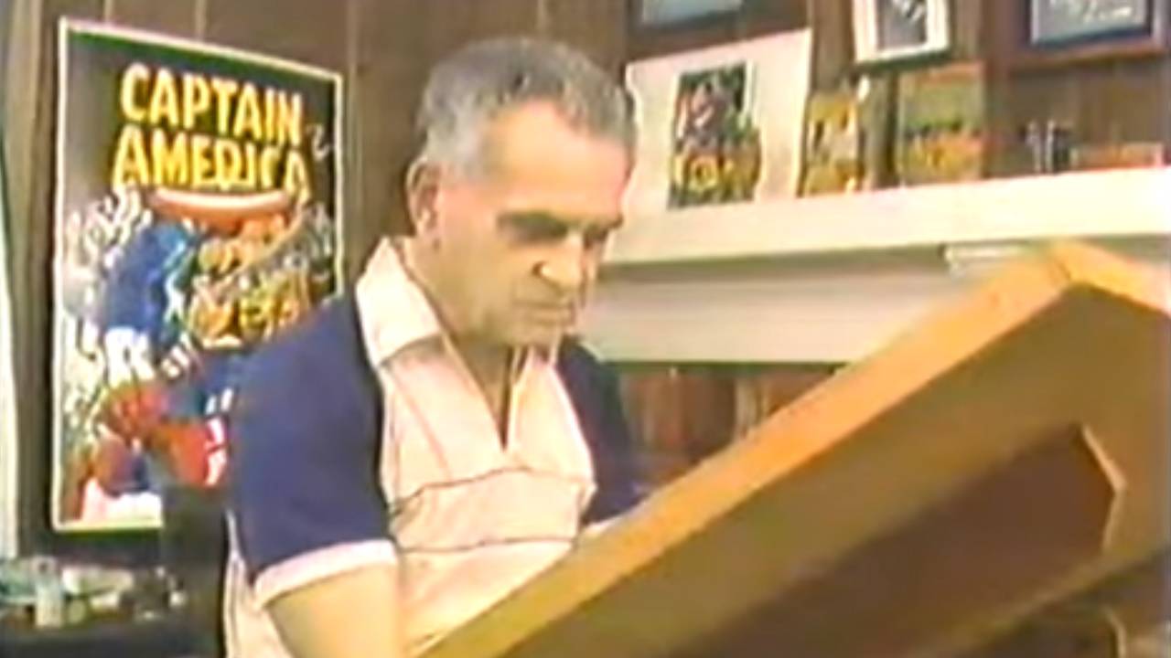 Jack Kirby sits at his drawing table, a Captain America poster behind him