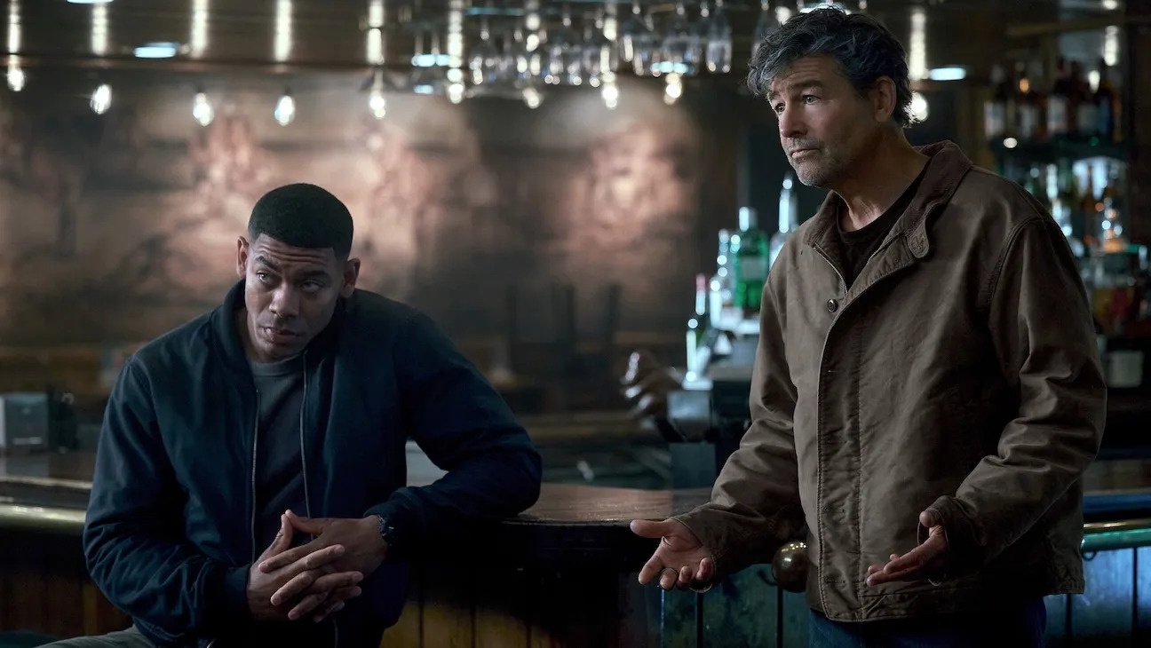 Two men, one seated and one standing, appear to be watching something in a bar