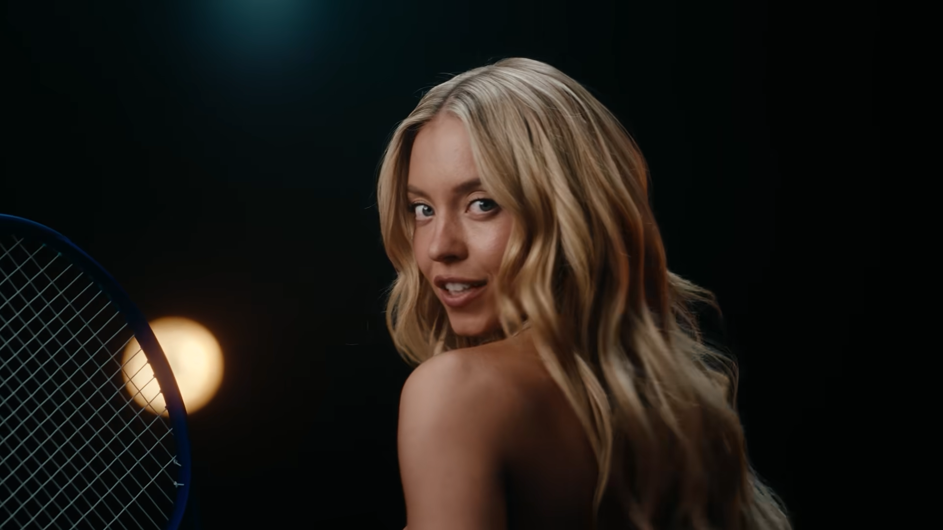 Sydney Sweeney holding a tennis racket and smiling over her bare shoulder