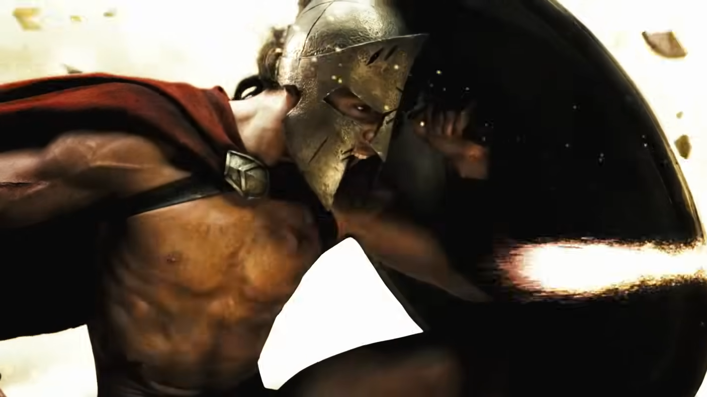 A shirtless Spartan warrior lifts his shield