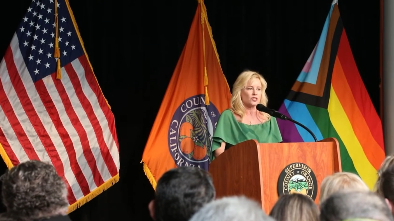 Woman behind podium and audience, with American flag, Orange County flag, and pride flag in background