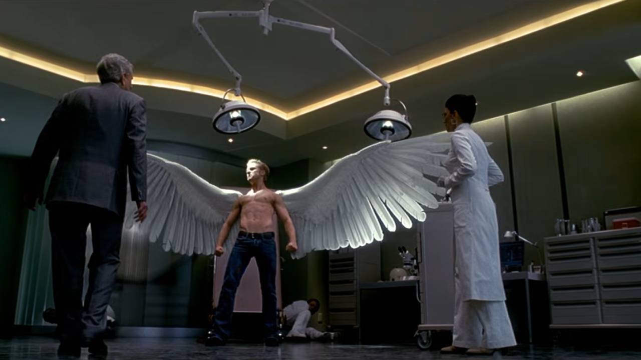 A shirtless young man with angel wings stands in a lab