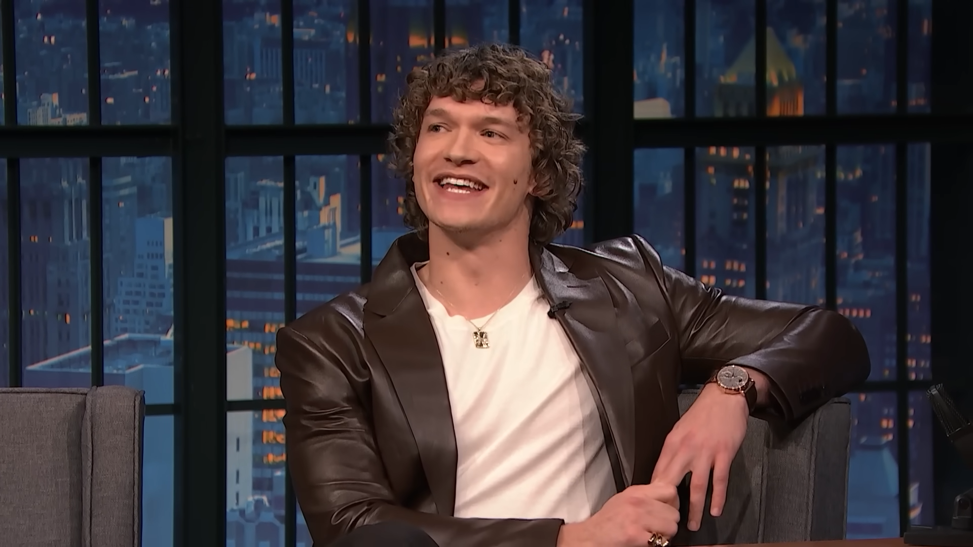 A young man smiles while seated for a late night show interview