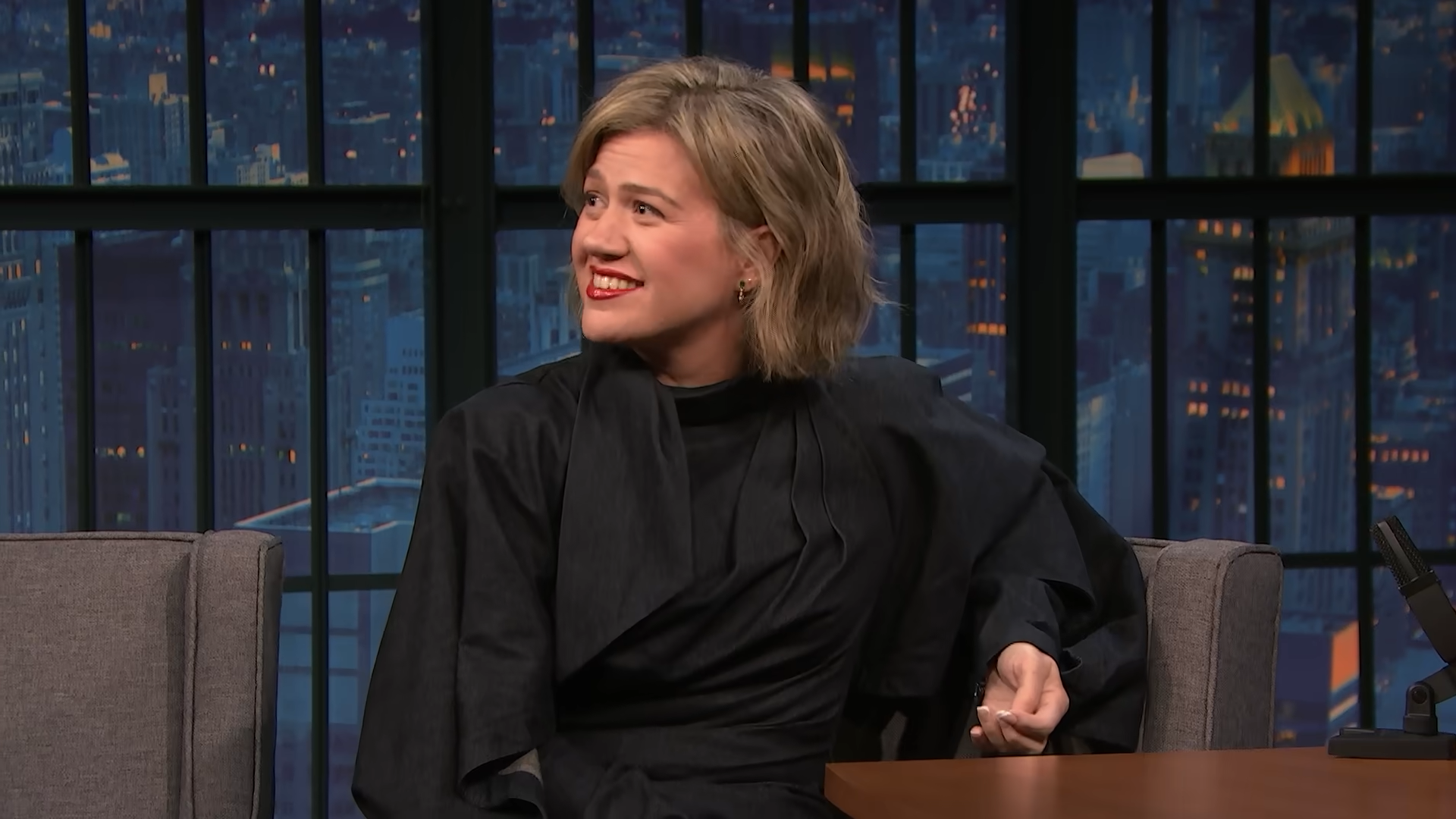 Kelly Clarkson seated for a late-night interview, giving a wincing smile