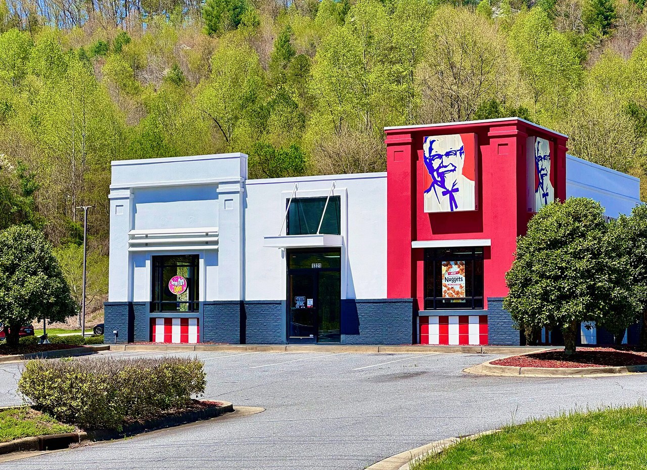 A red and white KFC restaurant with a Colonel Sanders mural, surrounded by green hills