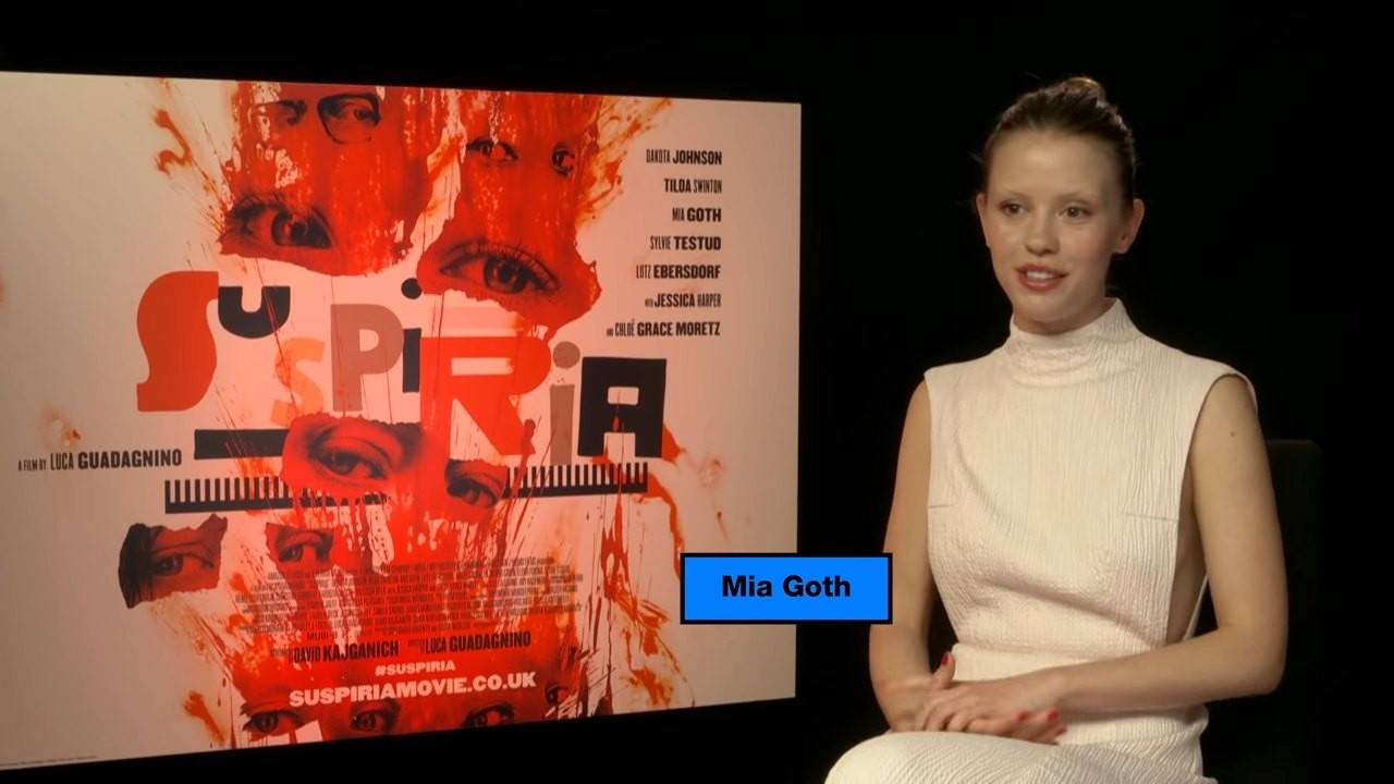 Mia Goth in a white sleeveless top seated in front of a red Suspiria poster during an interview
