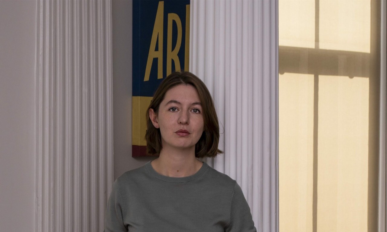 Sally Rooney in a gray sweater standing in a bright room with tall windows