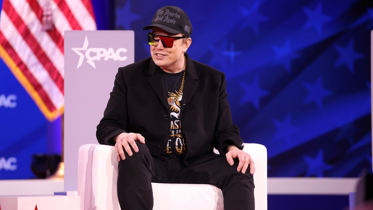 Elon Musk at CPAC 2025 featured 1280×720 Elon Musk wearing a black cap and sunglasses, seated on a white chair on a conference stage