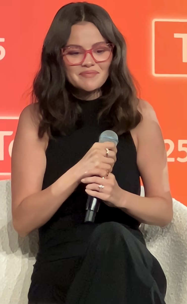 Selena Gomez in glasses and a black outfit speaking into a microphone on stage