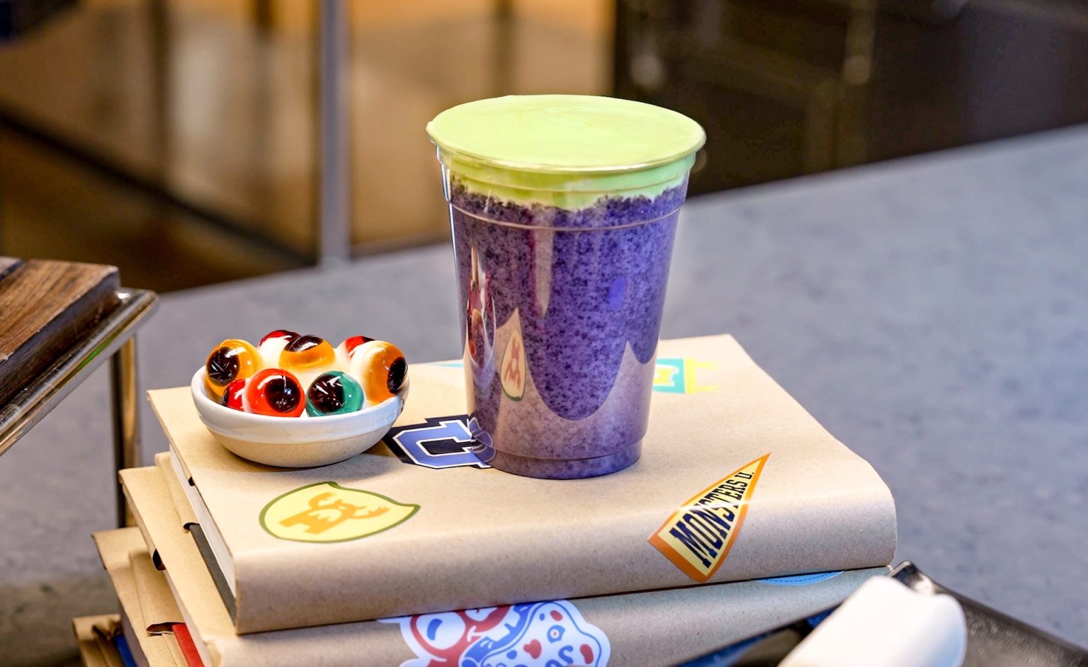 A purple frozen drink with green foam topping beside a dish of candy eyeballs on books covered in Monsters University stickers